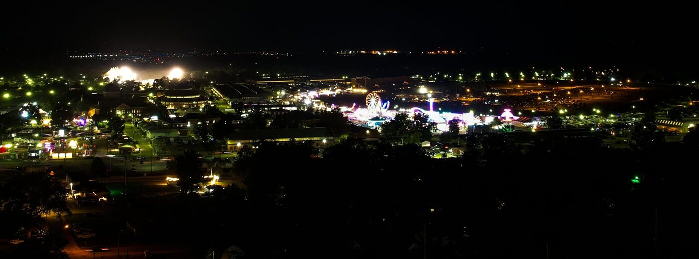 A nighttime aerial view of the Illinois State Fairgrounds lit up with midway rides, fair buildings, and parking areas.